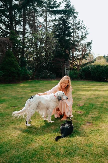 A woman in a garden smiles as she interacts with her dog and cat under lush trees.